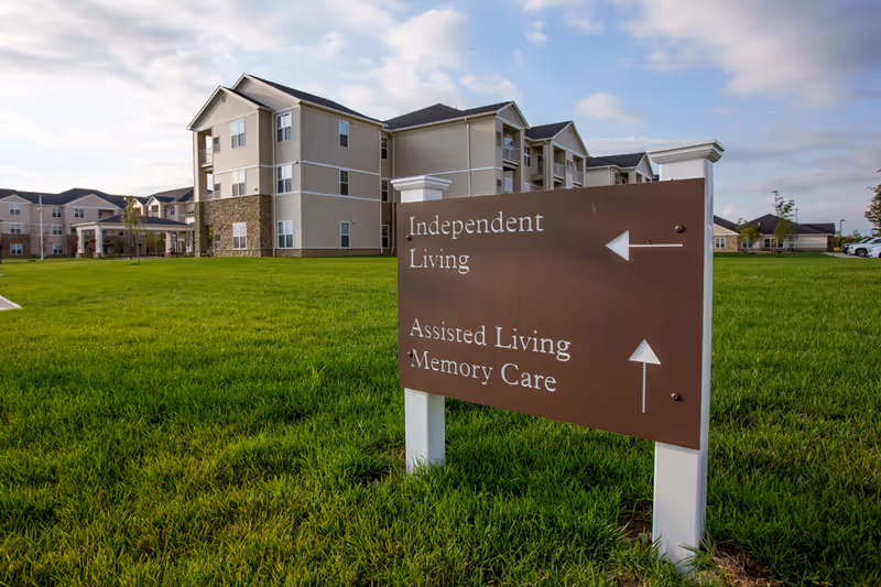 Outdoor view of a senior living facility with a large brown directional sign on a grassy lawn. The sign points left for Independent Living and straight ahead for Assisted Living and Memory Care. The multi-story building with beige siding and stone accents is visible in the background under a partly cloudy sky.