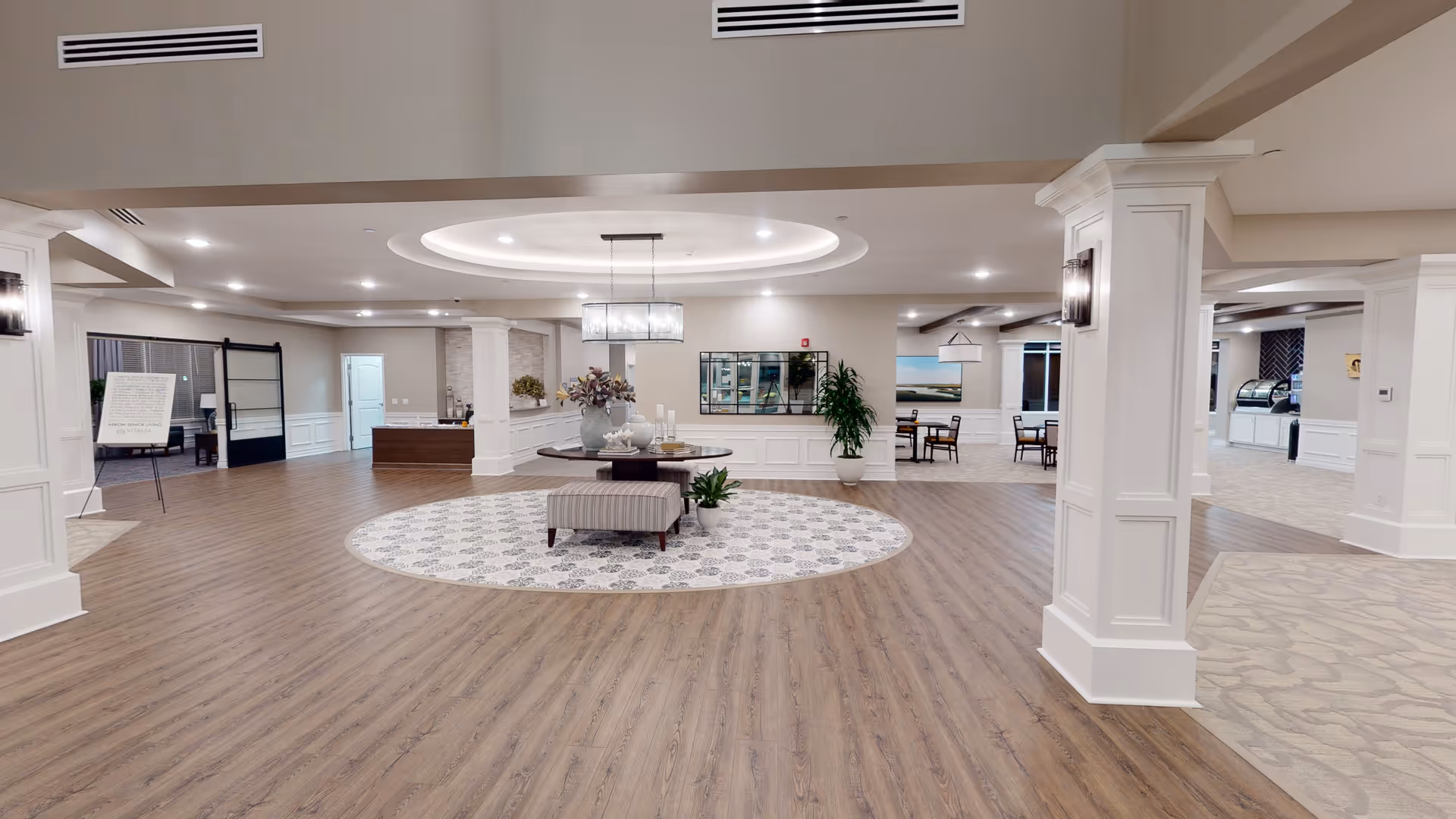 Spacious and well-lit interior common area of a senior living facility with wood flooring and a circular patterned rug in the center. A round table with decorative items and plants sits on the rug. The room features white columns, recessed lighting, and a modern chandelier. In the background, there are seating areas with tables and chairs, a reception desk, and a sign on an easel.