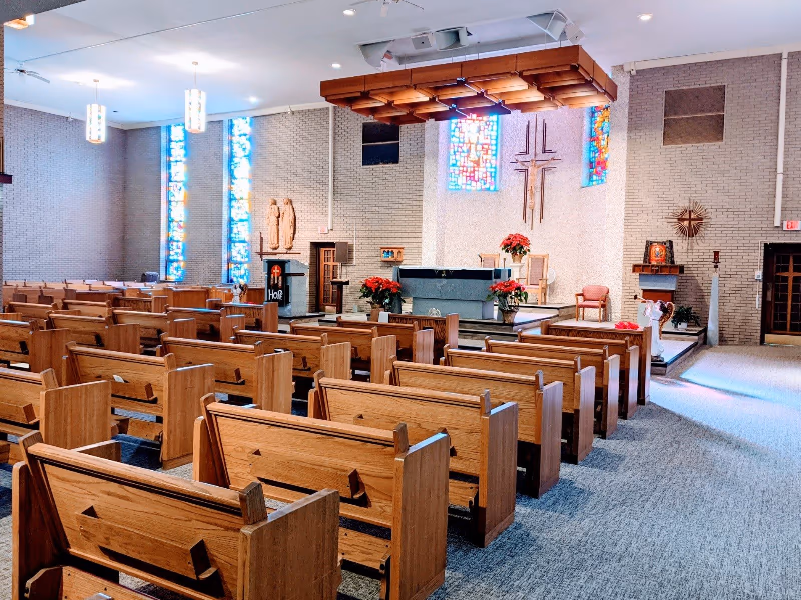 Interior view of a chapel with wooden pews arranged in rows facing an altar. The altar area features a large cross on the wall, stained glass windows, floral arrangements, and religious statues. The space is well-lit with ceiling lights and natural light from the stained glass windows.