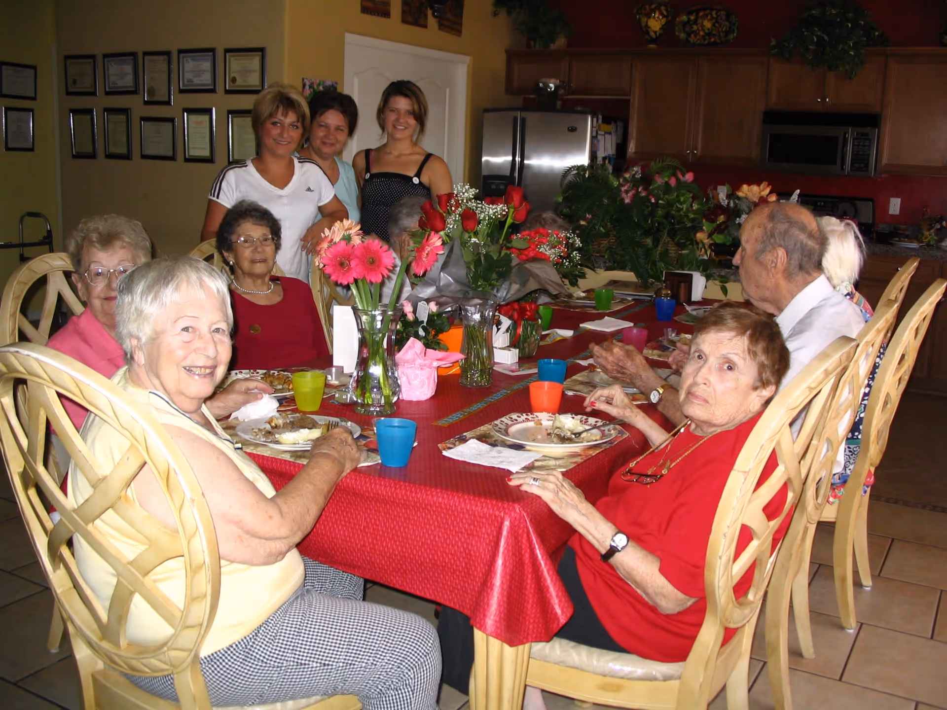 A group of elderly people sitting around a dining table covered with a red tablecloth, enjoying a meal together. There are three younger women standing behind them, smiling. The room has a kitchen area with wooden cabinets, a refrigerator, and a microwave in the background. The table is decorated with vases of flowers and colorful cups.