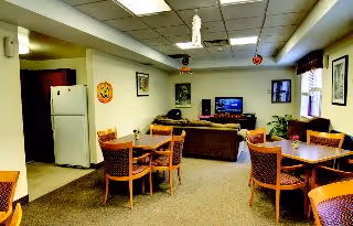 A common area in Heritage Pointe Apartments featuring multiple wooden tables with chairs, a brown couch facing a television, a white refrigerator in an open kitchen area, and ceiling decorations including hanging ornaments.