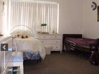 A small bedroom with two beds, one white metal-framed bed with stuffed animals and a floral bed skirt, and one hospital-style bed with a purple blanket. There is a white dresser with a green lamp and a clock on top, a white stool, and vertical blinds covering the window.