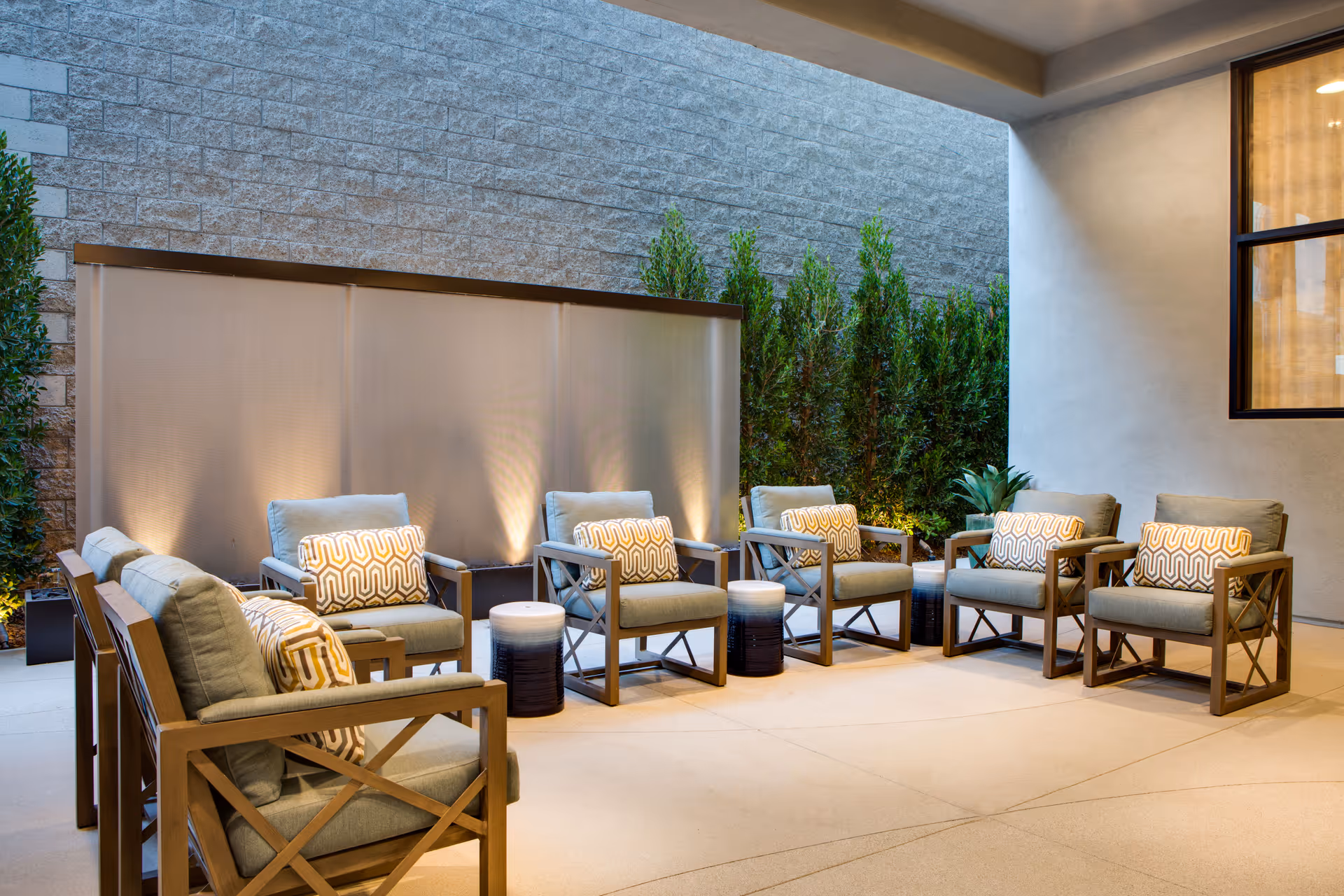 A modern outdoor seating area with eight cushioned armchairs arranged in a circle around small round tables. The space is enclosed by a stone wall and a privacy screen with soft lighting, and there are tall green plants along the back wall.
