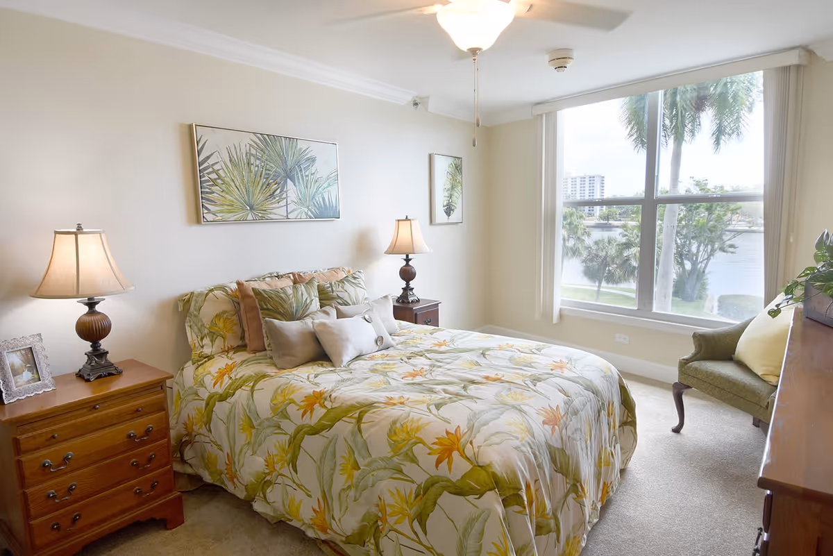 A bright bedroom with a large window showing a view of palm trees and water outside. The room features a bed with floral bedding and multiple pillows, two wooden nightstands with lamps, framed botanical artwork on the walls, a green upholstered chair with a yellow pillow, and a wooden dresser with a plant on top.