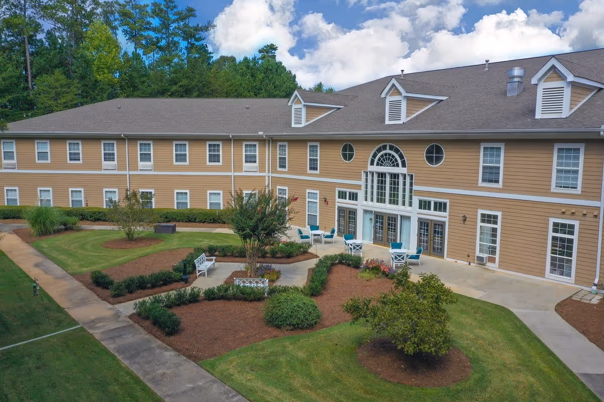 Exterior view of a two-story senior living facility building with beige siding and white trim. The building features multiple windows and a large central entrance with tall windows and decorative circular windows above. In front of the building is a landscaped garden area with trimmed bushes, small trees, flower beds, and white benches along paved walkways. The sky is partly cloudy with blue patches visible.