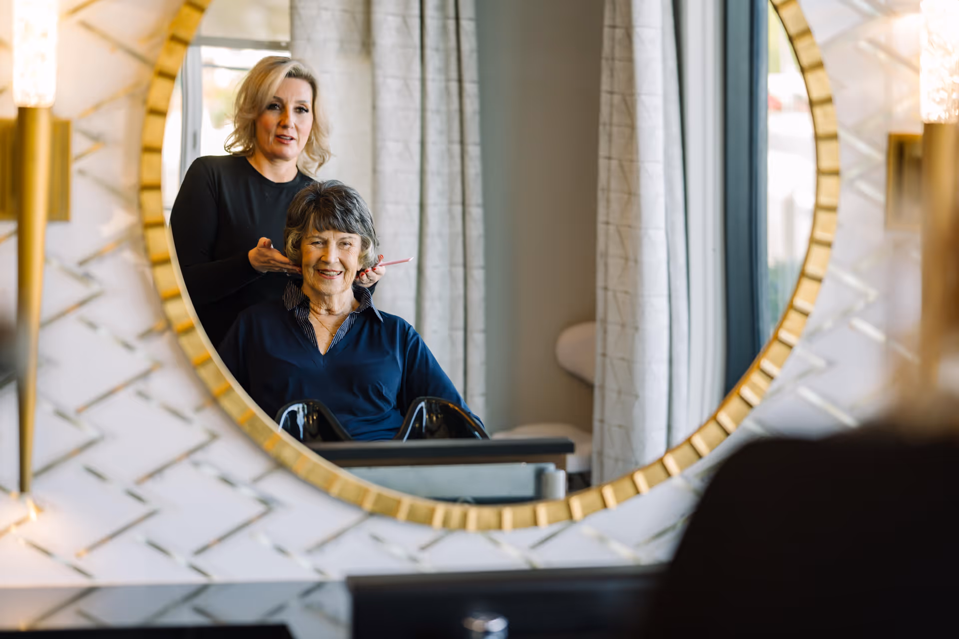An elderly woman sitting in a chair smiling while a hairstylist stands behind her, styling her hair. They are reflected in a large round mirror with a gold frame, in a well-lit room with patterned white walls and curtains.