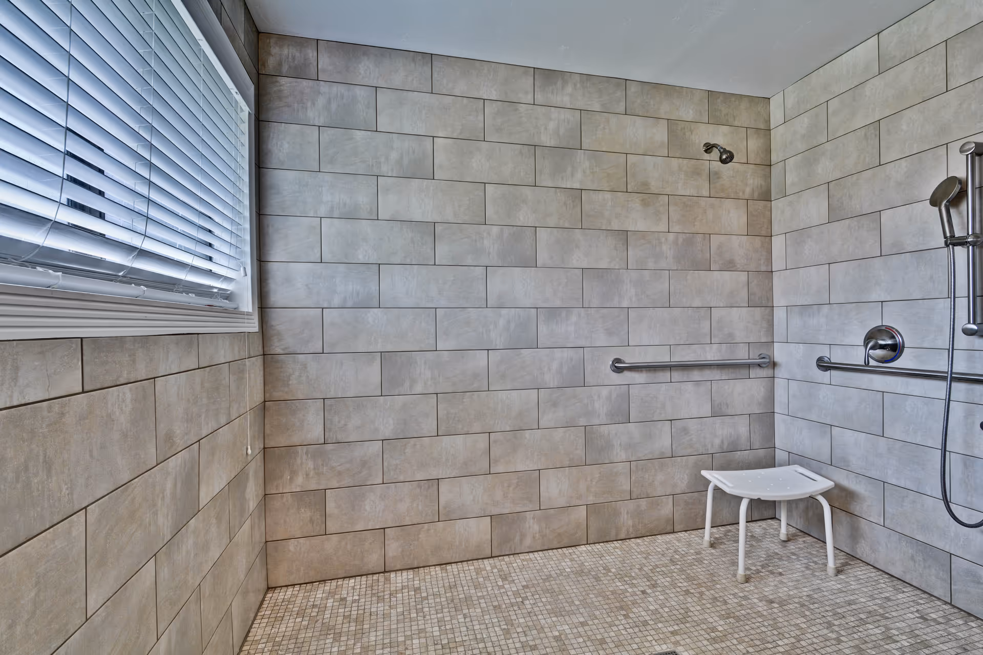 A tiled walk-in shower with beige rectangular wall tiles and small square floor tiles. The shower features a fixed showerhead, a handheld showerhead on a sliding bar, two horizontal grab bars, and a white shower stool. A window with white blinds is visible on the left wall.