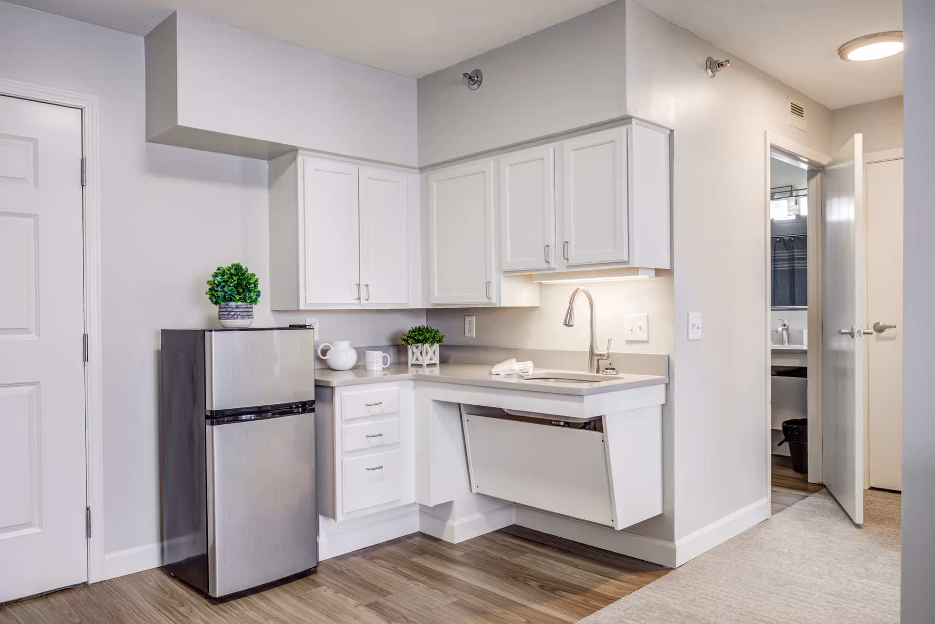 Small modern kitchenette with a stainless steel mini fridge, white cabinets, countertop sink, and an open door leading to a bathroom.