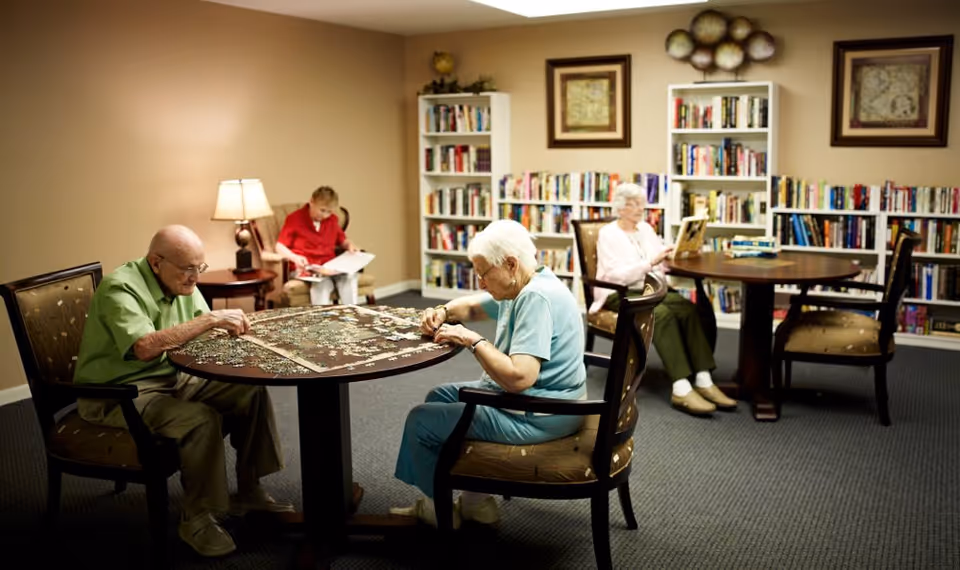 A cozy room in a senior living facility with four elderly individuals engaged in quiet activities. Two seniors are seated at a round table working on a large jigsaw puzzle, while two others are seated separately reading books. The room has bookshelves filled with books, framed artwork on the walls, and soft lighting from a table lamp.