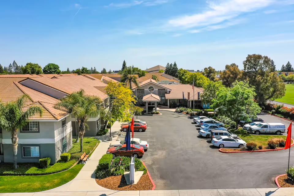 Aerial view of Fresno Senior Living facility showing a large parking lot with several parked cars, surrounded by buildings with tan roofs and gray walls. The area is landscaped with palm trees, bushes, and green grass under a clear blue sky.
