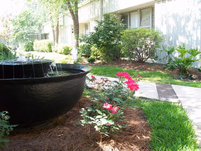 Outdoor garden area with a large black water fountain, blooming pink flowers, green bushes, and a concrete walkway next to a building with windows.
