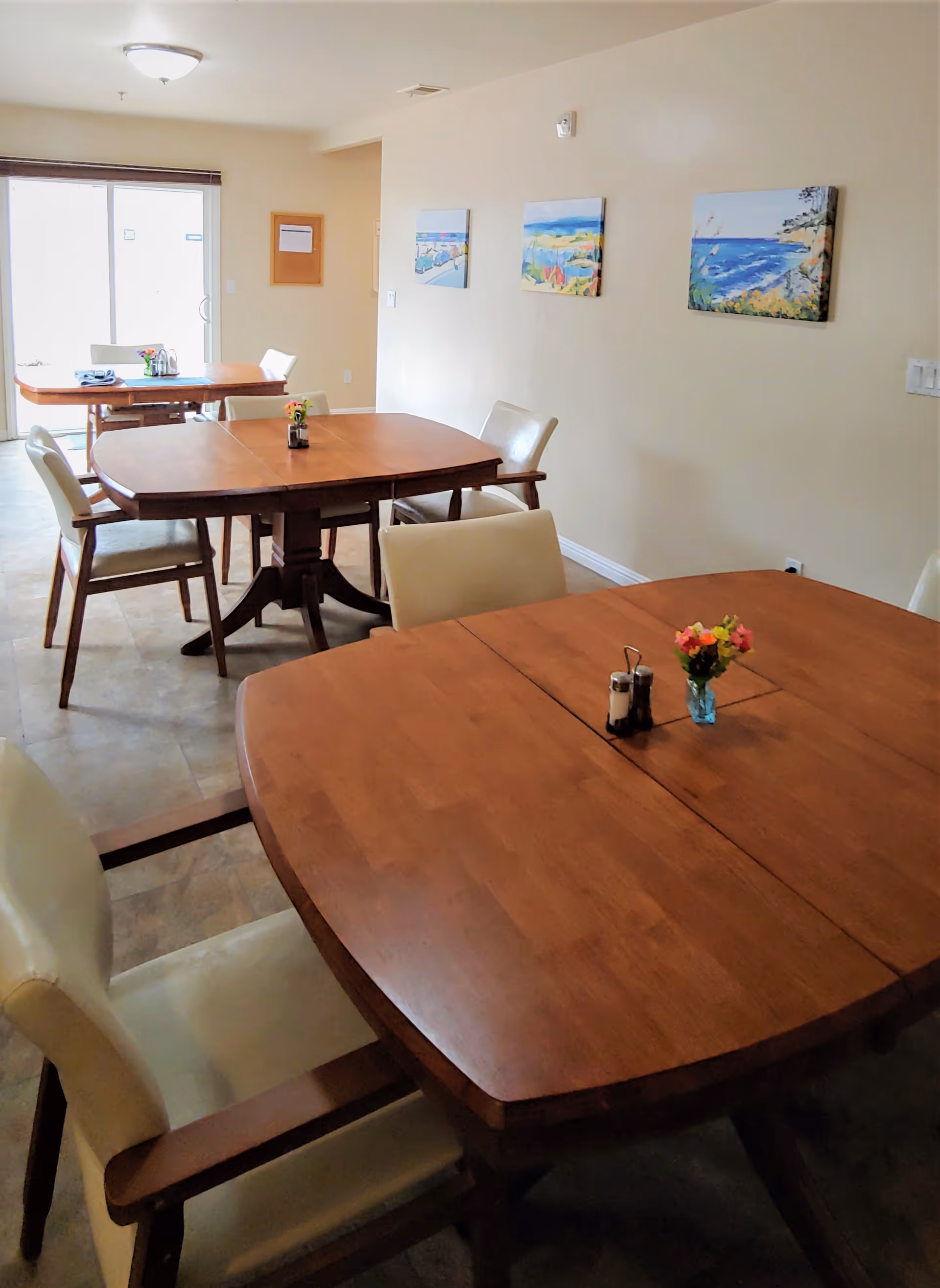 A dining room with several wooden tables and cream-colored cushioned chairs. Each table has a small vase with flowers and a salt and pepper shaker set. The room has beige walls adorned with three colorful landscape paintings. A sliding glass door is visible in the background, allowing natural light to enter.