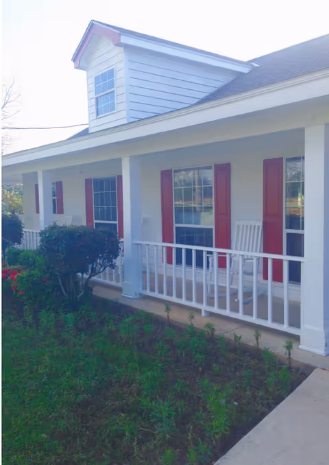 White porch with rocking chairs along the front of a light-blue building with red shutters.