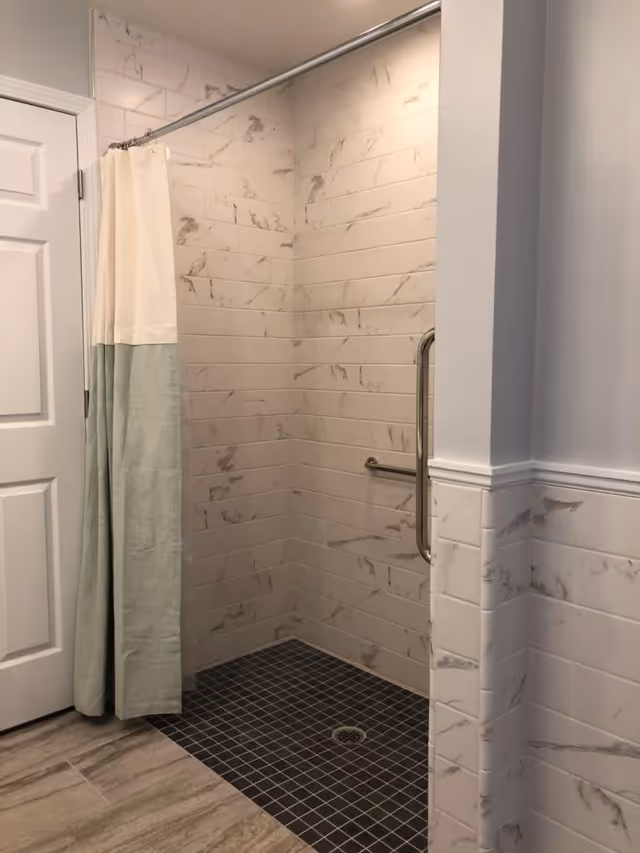 Walk-in tiled shower with marble-look subway tile walls, dark square floor tiles, a grab bar, and a two-tone shower curtain.