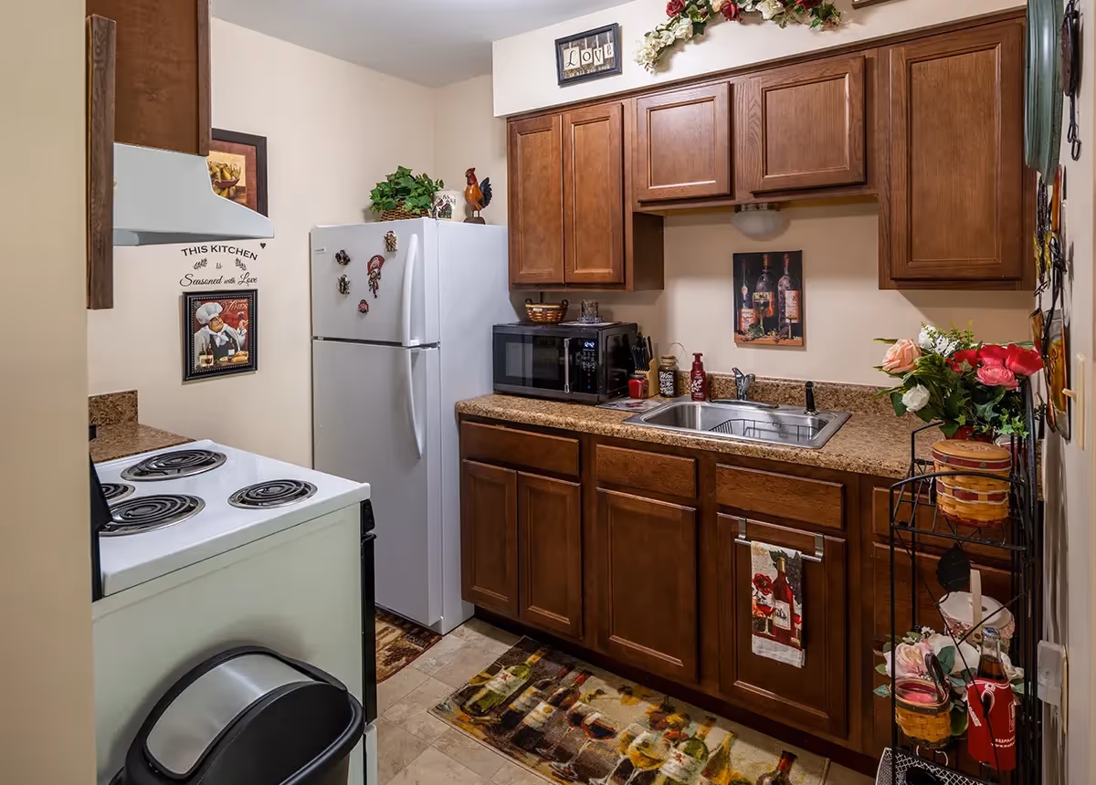 Small kitchen with wooden cabinets, a white refrigerator, a white electric stove, a microwave on the countertop, a stainless steel sink, and various decorative items including flowers, a wall painting, and kitchen-themed signs.