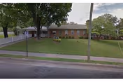 Single-story brick building with a large front lawn and several trees, viewed from across the street. The building appears to be a residential or assisted living facility with a white fence on the left side and a sidewalk in front.