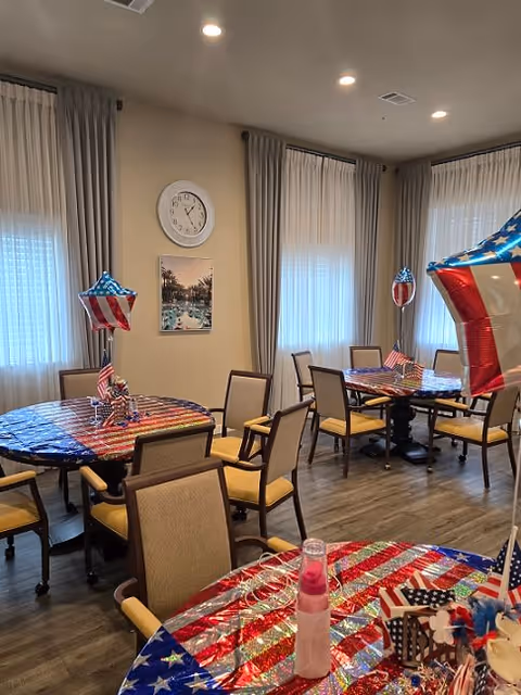 Communal dining room with round tables and chairs decorated with American flag–themed tablecloths, small flags and balloons.
