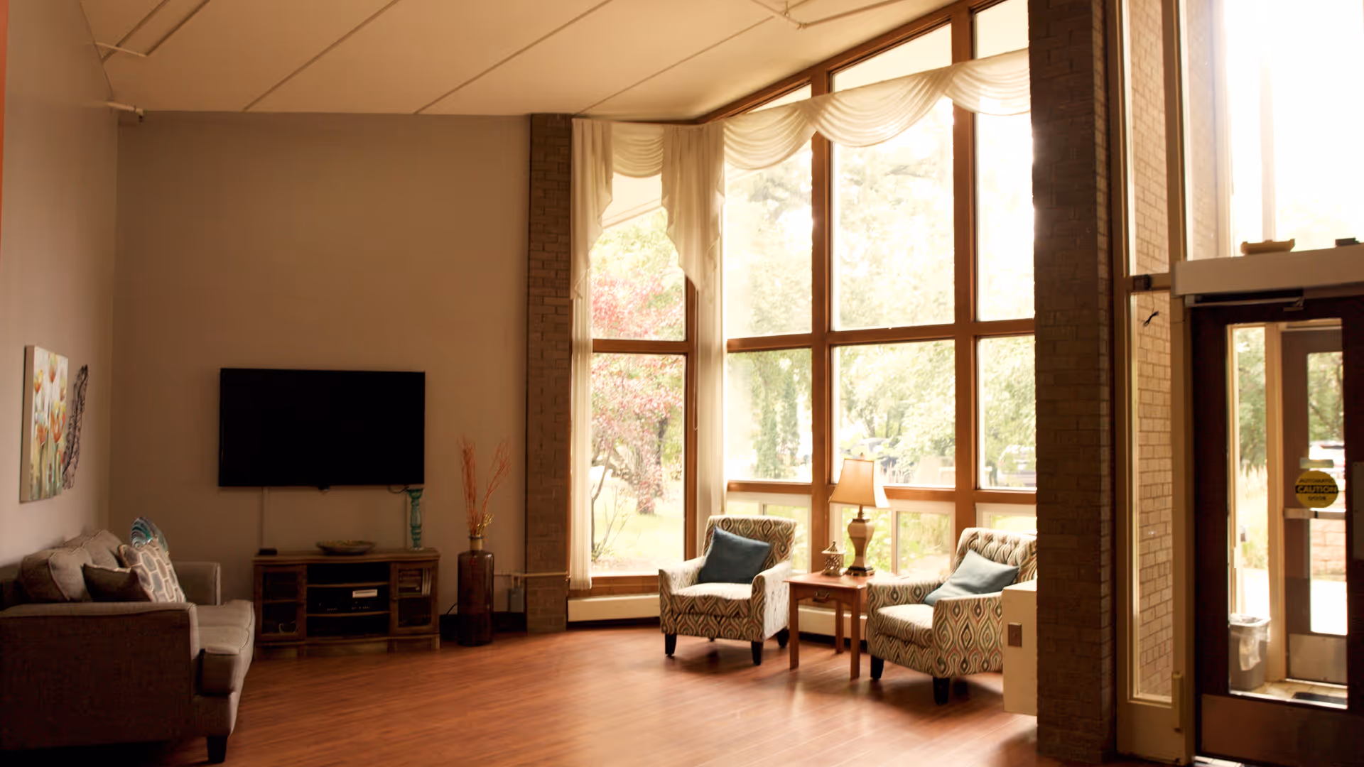 A bright living room area with large floor-to-ceiling windows letting in natural light. The room features a gray sofa with patterned pillows, a wall-mounted flat-screen TV above a wooden media console, two patterned armchairs with blue cushions, a wooden side table with a lamp, and a glass door leading outside.