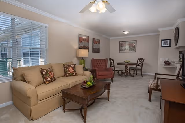 A cozy living room area featuring a beige sofa with floral cushions, a wooden coffee table with a small plant centerpiece, a red armchair, and a small round dining table with two chairs in the background. The room has beige walls, a ceiling fan with lights, a window with white blinds, and framed artwork on the walls.