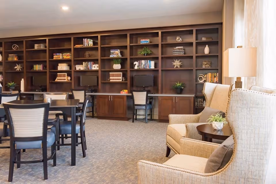 A cozy common area with beige armchairs and a small round table with a plant, adjacent to a dark wood bookshelf filled with books and decorative items. There are several desks with chairs and computer monitors along the bookshelf. The room is well-lit with natural light coming through sheer curtains.