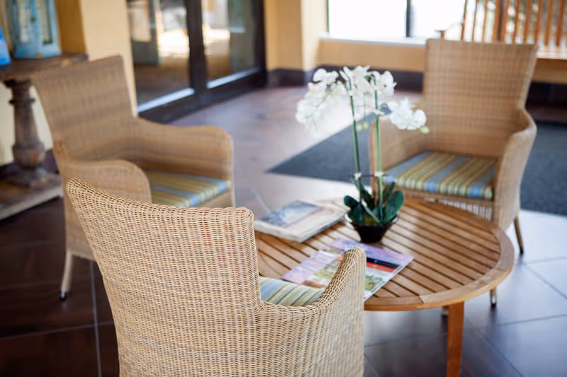 A cozy seating area with three wicker chairs featuring striped cushions arranged around a round wooden table. On the table, there is a small potted white orchid and some magazines. The setting appears to be indoors near large windows and a glass door.