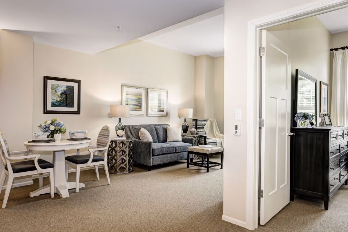A cozy living room and dining area in a senior living facility. The living room features a gray sofa with cushions, two side tables with lamps, and a small coffee table. The dining area has a round white table with four chairs, set with plates and a flower centerpiece. The walls are decorated with framed artwork. To the right, an open door reveals a bedroom with a dark dresser, a mirror, and a window with curtains.