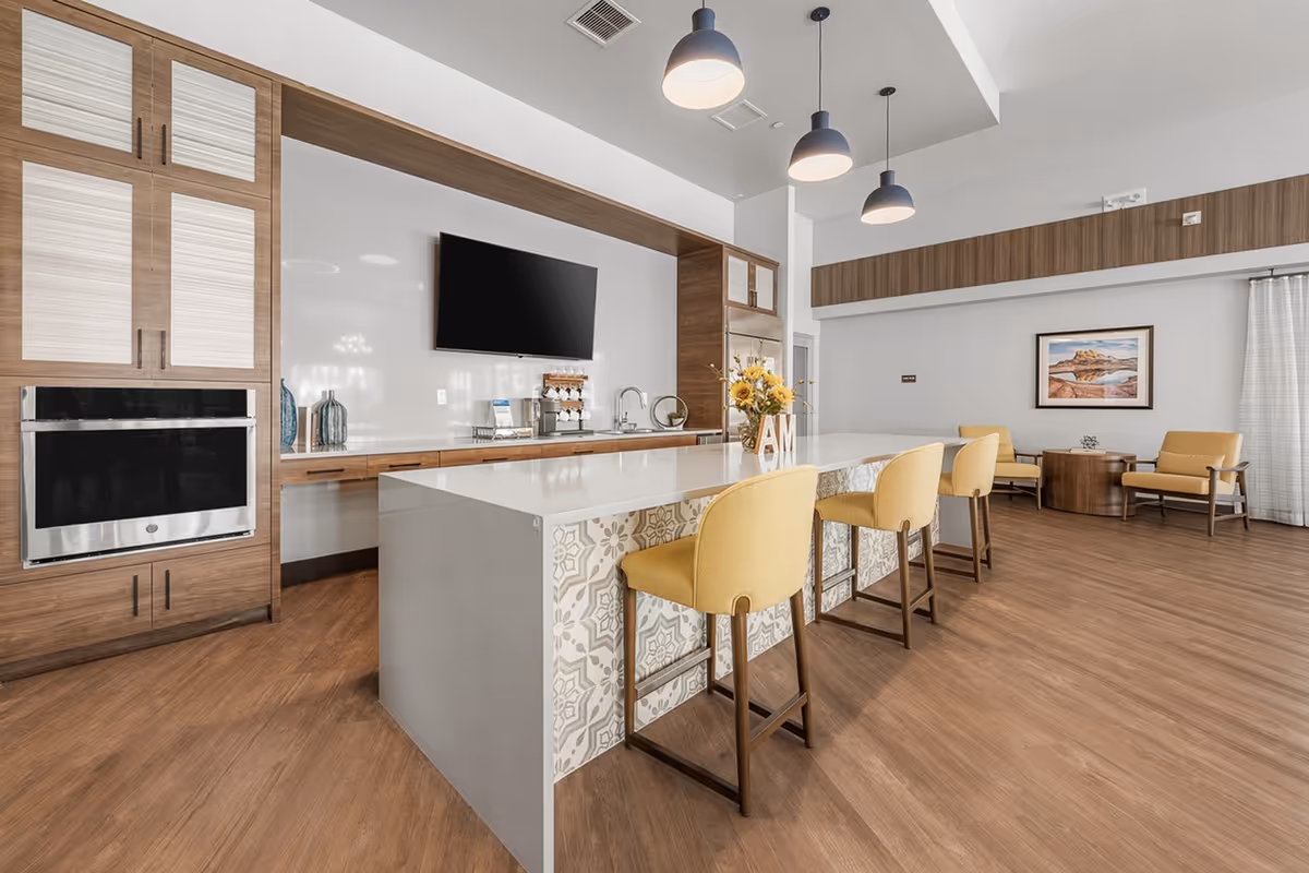 Modern senior living facility kitchen and lounge area with a large island featuring patterned tiles and four yellow cushioned bar stools. The kitchen has wooden cabinetry, a built-in oven, a mounted flat-screen TV, and a coffee station. In the background, there is a seating area with two yellow armchairs, a round wooden table, a framed landscape picture on the wall, and large windows with white curtains.
