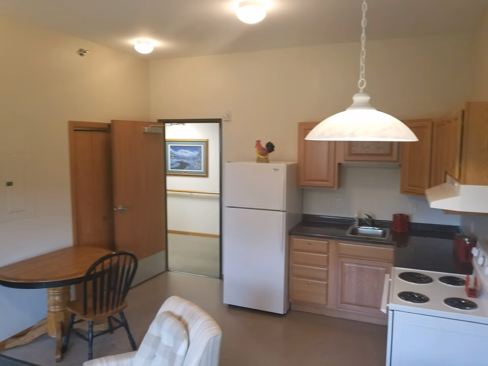 Interior view of a small kitchen area in a retirement community unit, featuring wooden cabinets, a white refrigerator, an electric stove, a sink, a wooden table with a single chair, and a cushioned armchair. The room is lit by ceiling lights and a hanging lamp above the kitchen counter. An open door leads to a hallway with a framed picture on the wall.