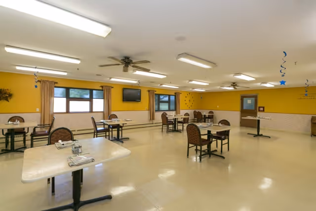 A spacious dining room with several tables and chairs arranged neatly. The walls are painted yellow with a beige lower half, and there are windows with curtains along one wall. Ceiling fans and fluorescent lights are mounted on the ceiling. A flat-screen TV is mounted on the wall, and some decorative elements are visible on the walls.