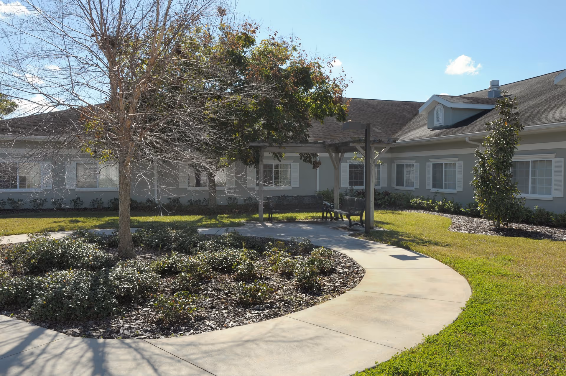 Outdoor courtyard area at Life Care Center of Winter Haven featuring a curved concrete walkway, landscaped garden beds with shrubs and trees, benches under a wooden pergola, and a building with multiple windows in the background.