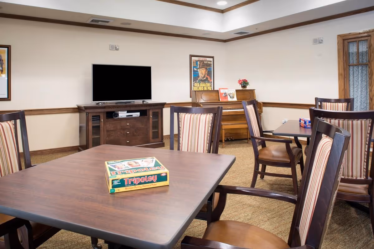 Community activity room with tables and striped chairs, a TV on a console, and a piano in the background.
