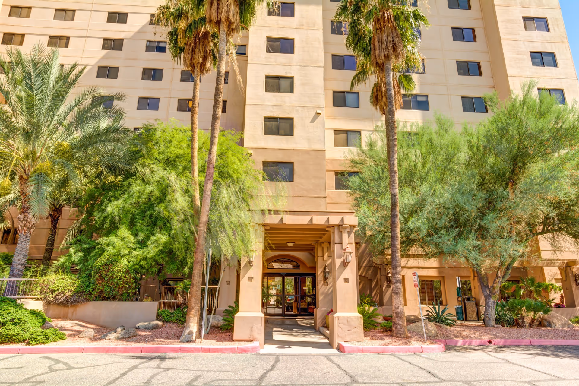 Exterior view of Courtyard Towers, a multi-story beige building with several windows. The entrance is framed by a covered walkway supported by columns. Tall palm trees and green shrubs surround the entrance area, with a paved driveway in front.