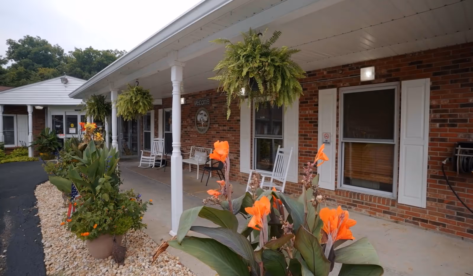 Front porch area of a brick building with white pillars and hanging green plants. There are orange flowers in pots along the walkway, white rocking chairs, and a welcome sign on the wall. The building has white window shutters and a covered walkway.