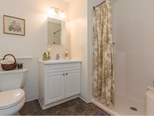 A clean bathroom featuring a white toilet with a basket on top, a white vanity with a sink and mirror above it, a wall-mounted light fixture, and a shower area with a floral-patterned shower curtain.