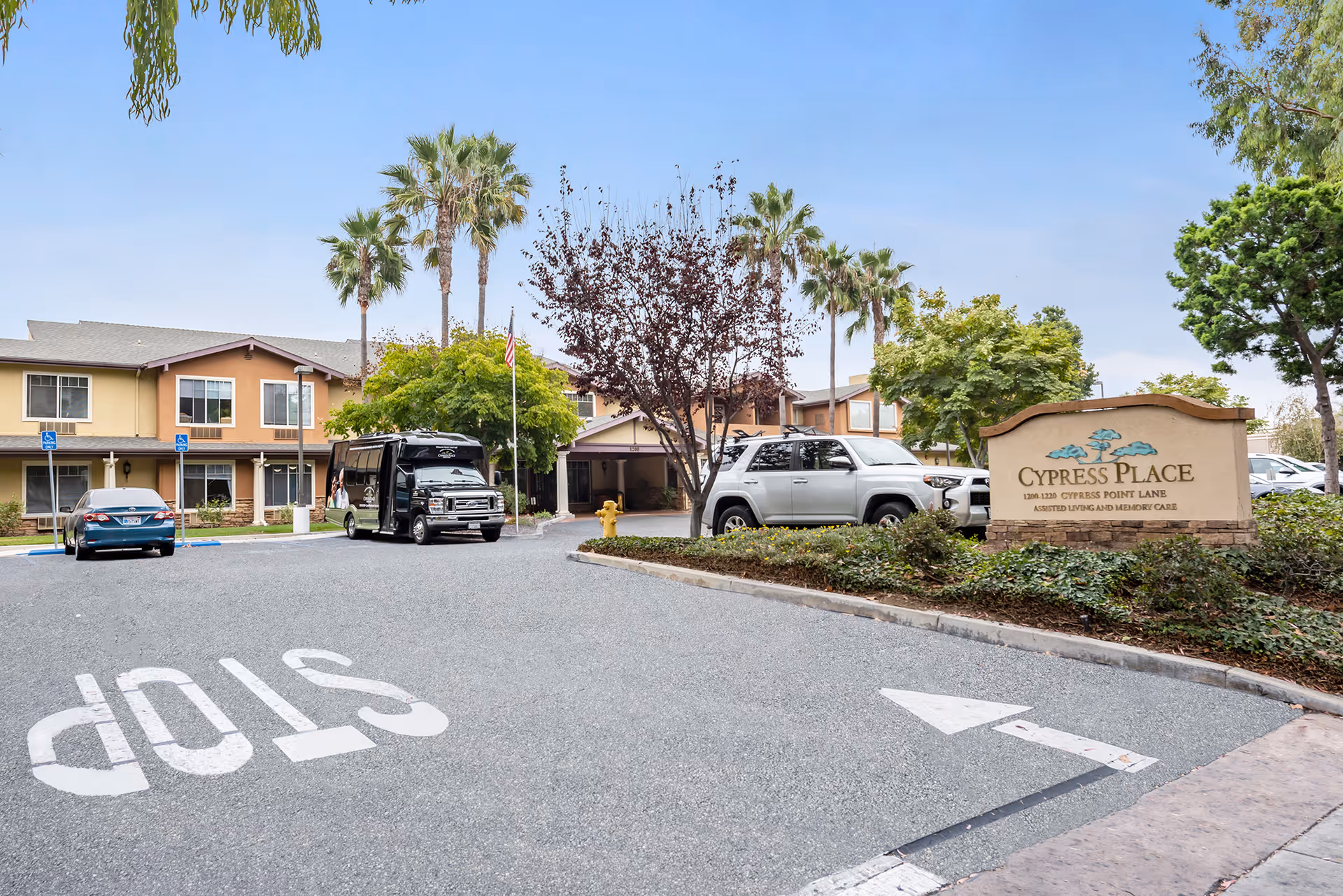 Exterior view of Cypress Place assisted living and memory care facility with a driveway, parked cars, palm trees, and a sign displaying the facility name and address.