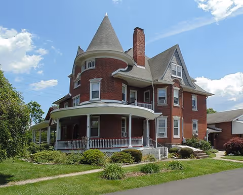 Large, historic red brick house with a conical turret, multiple chimneys, and a wraparound porch surrounded by green lawn and shrubs under a partly cloudy blue sky.