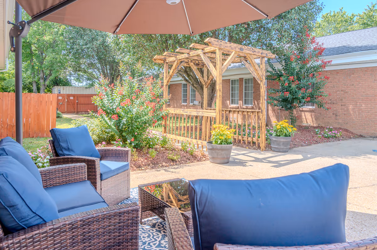 Outdoor patio with wicker seating and blue cushions under an umbrella facing a wooden pergola, potted plants, and a brick building.