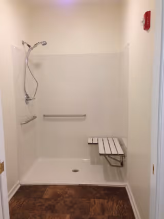 A walk-in shower with a handheld showerhead mounted on the left wall, two horizontal grab bars, and a foldable white bench attached to the right wall. The shower has a white interior and a brown floor outside the shower area.