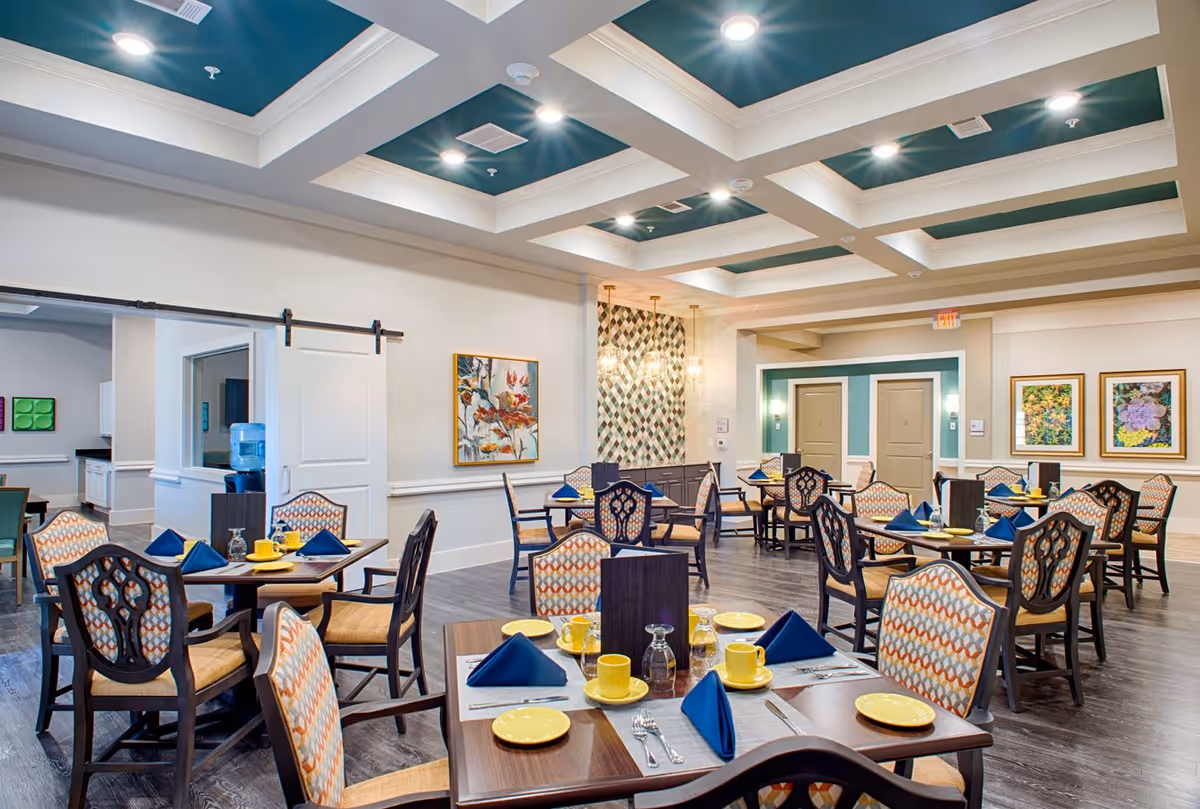 A bright and spacious dining room in an assisted living facility with multiple wooden tables set with yellow plates, cups, and blue napkins. The room features patterned upholstered chairs, a coffered ceiling with recessed lighting, decorative wall art, and a sliding barn door leading to another area.