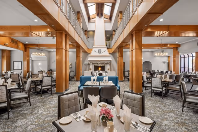 Spacious dining room in a senior living community with multiple tables set for meals, featuring folded white napkins, plates, cups, and silverware. The room has wooden beams, a high ceiling with skylights, a large fireplace in the center, and comfortable chairs arranged around the tables. The carpet has a patterned design, and there are chandeliers providing lighting.