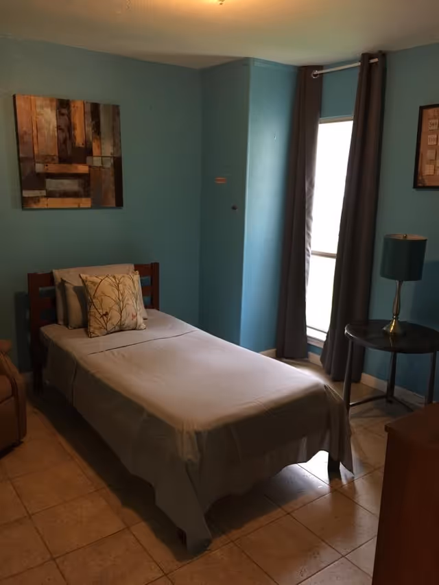 A small bedroom with a single bed covered in a light gray sheet and two pillows, one with a floral pattern. The walls are painted blue, and there is a window with gray curtains letting in natural light. A round side table with a green lamp is next to the bed, and a piece of abstract art hangs on the wall above the bed.