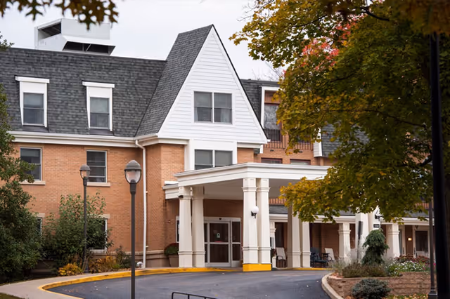 Front entrance of a brick senior living building with a white columned porte-cochere and surrounding trees.
