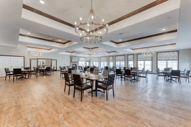 Spacious dining room in a senior living facility with multiple tables and chairs arranged neatly. The room features large windows with patterned blinds, wooden flooring, and elegant chandeliers hanging from a coffered ceiling with wooden accents.