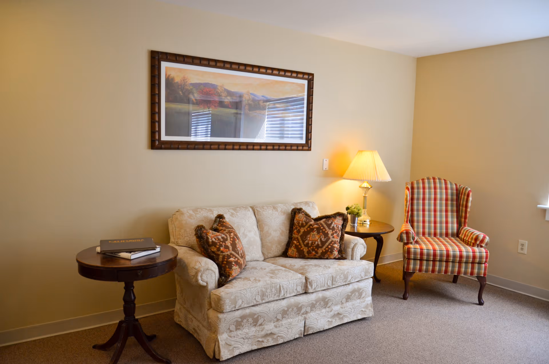 A cozy sitting area with a patterned loveseat, plaid armchair, side tables, a lamp, and framed wall art.