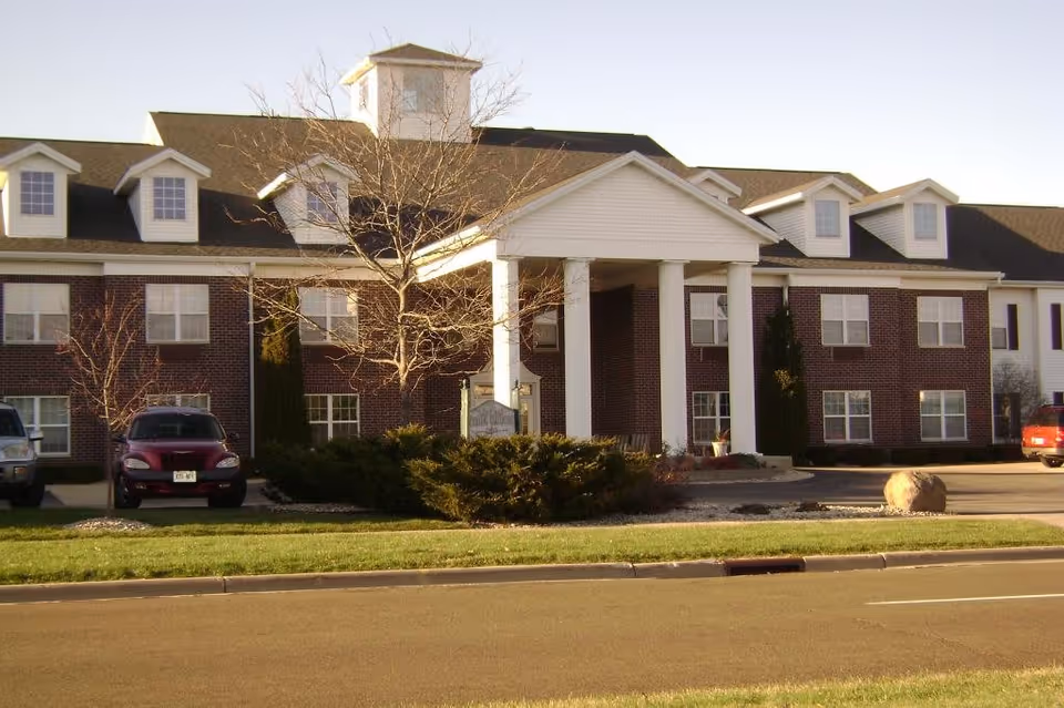 Exterior view of Prairie Gardens Assisted Living facility showing a two-story brick building with white columns at the entrance, several windows, a few parked cars, and a clear sky.