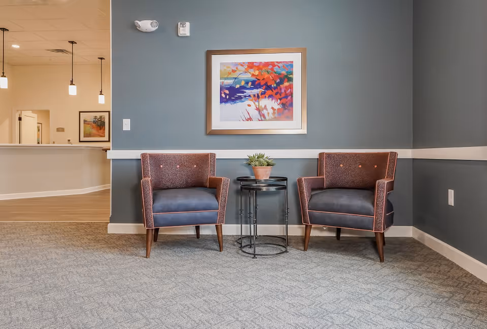 A seating area in a senior living facility with two upholstered armchairs separated by a small round black table holding a potted plant. The wall behind is painted gray with a white chair rail and features a colorful framed painting. The floor is carpeted, and the adjacent room has wood flooring and pendant lights hanging from the ceiling.