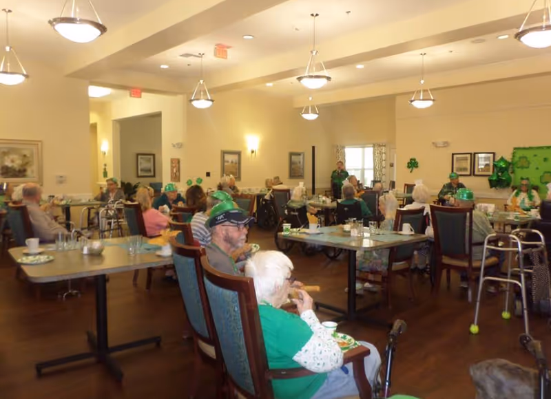 A dining room in a senior living facility with elderly residents seated at tables. The room is decorated with green shamrock decorations and balloons, suggesting a St. Patrick's Day celebration. The residents are engaged in eating and socializing, and some are wearing green hats. The room has warm lighting and framed artwork on the walls.