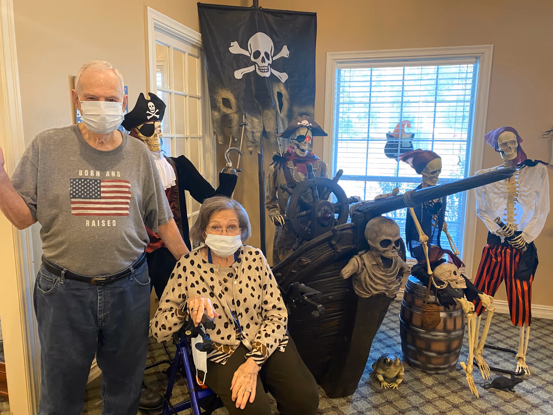 An elderly man and woman wearing face masks pose indoors next to a pirate-themed Halloween display featuring skeletons dressed as pirates, a pirate ship wheel, a cannon, and a pirate flag with a skull and crossbones. The woman is seated with a walker, and the man stands beside her.