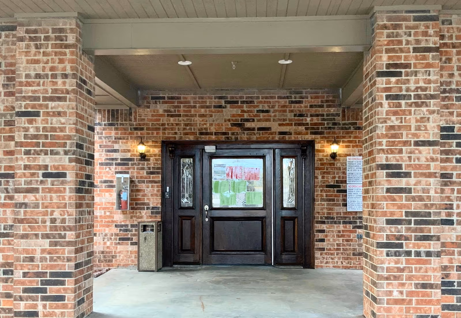 Entrance to a building with a dark wooden door framed by brick walls and two lit wall-mounted lanterns on either side. There is a fire extinguisher case and a trash can to the left of the door, and a sign with rules on the right brick wall. The area is covered by a ceiling with recessed lighting.