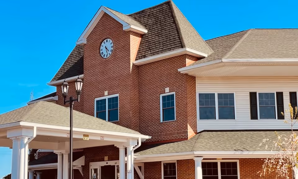 Exterior view of a senior living facility building with a brick facade and a clock tower. The building has multiple windows, a covered entrance supported by white columns, and a clear blue sky in the background.